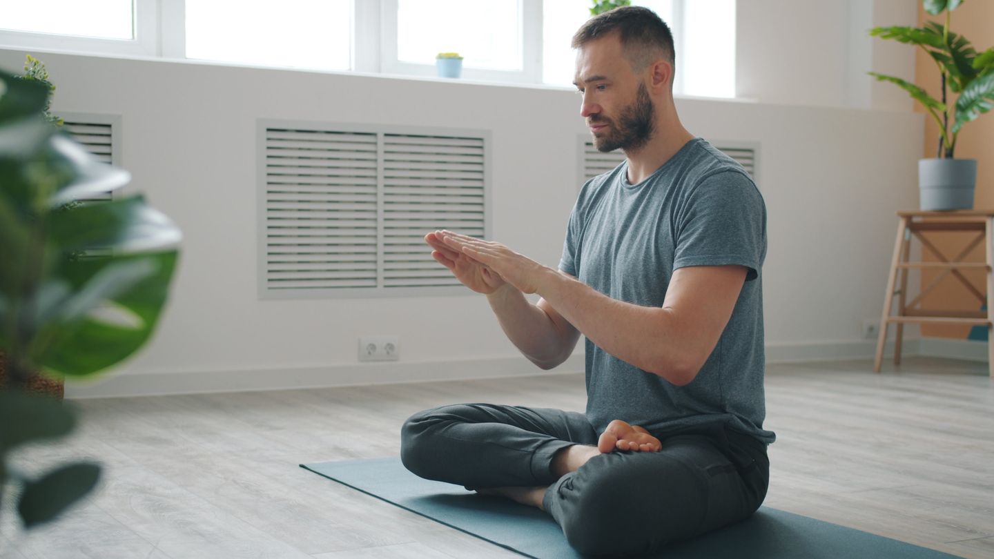 Adult man meditating on a yoga mat in bright indoor space, focusing on self-care and mindfulness.
