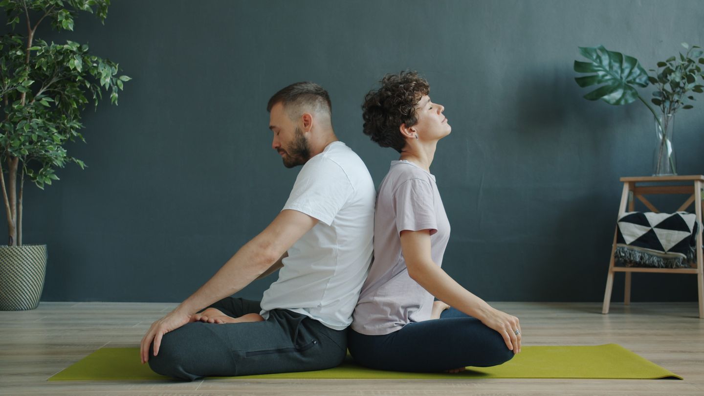 A couple meditating back to back on a yoga mat indoors, promoting relaxation and mindfulness.